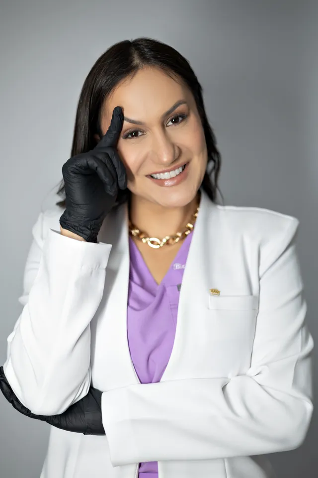 Bianca in a white coat and black gloves, touching her temple and smiling